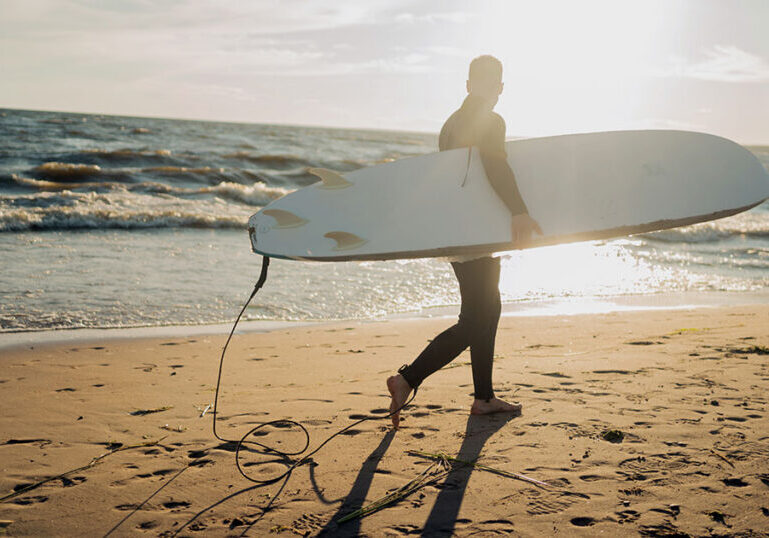Surfer carrying a surfboard along a New Zealand beach at sunset, representing the lifestyle of American expats living in New Zealand who must navigate the foreign investment fund tax regime
