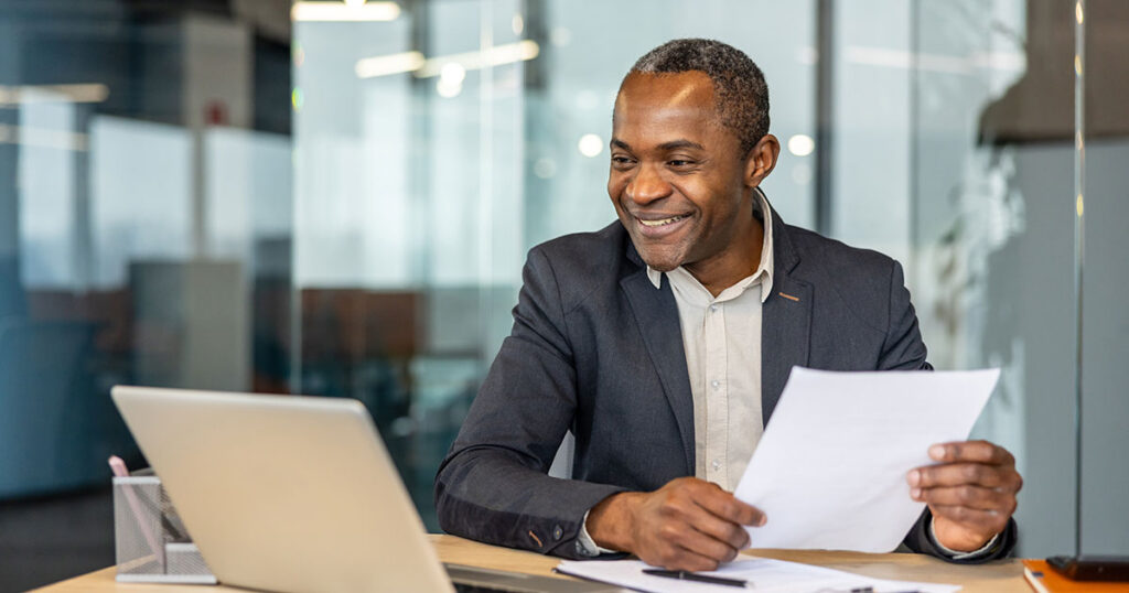 Corporate executive smiling while reviewing financial documents at a laptop, symbolizing comprehensive executive financial planning that aligns compensation, investments, taxes and cash flow with long‑term goals