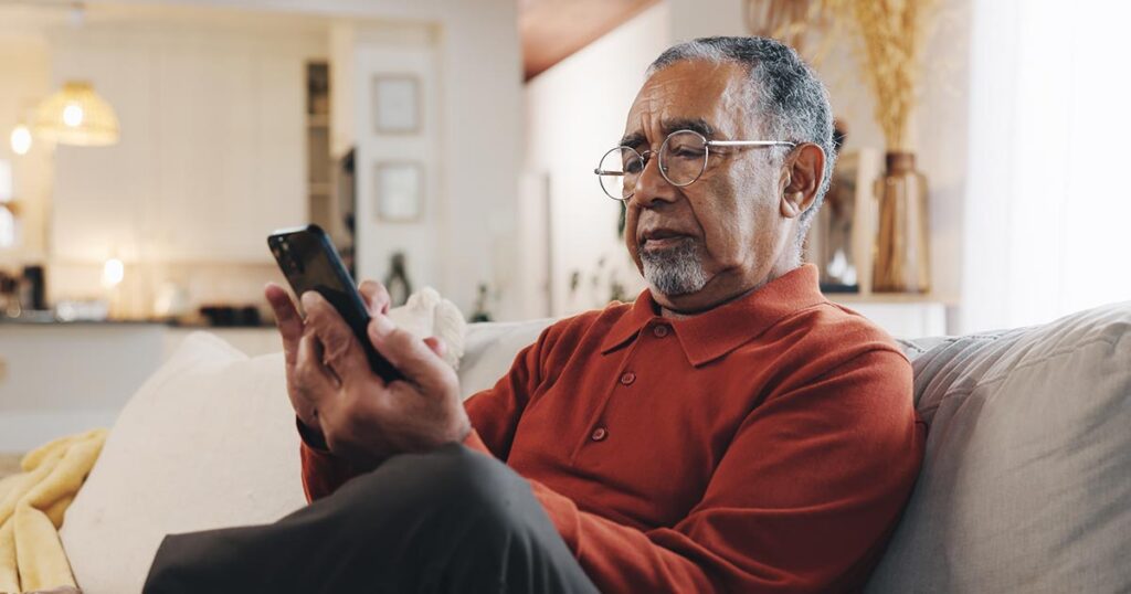 An older man sits on a living room couch, looking skeptically at his smartphone as he investigates a potential investment message, illustrating caution against online scams and financial fraud.