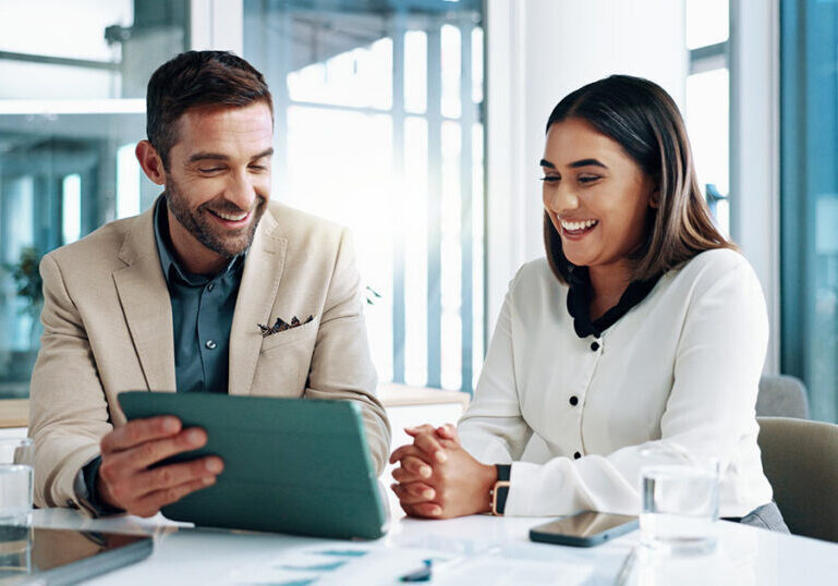 A male and female financial advisor smiling while reviewing wealth management strategies on a digital tablet in a modern corporate office.