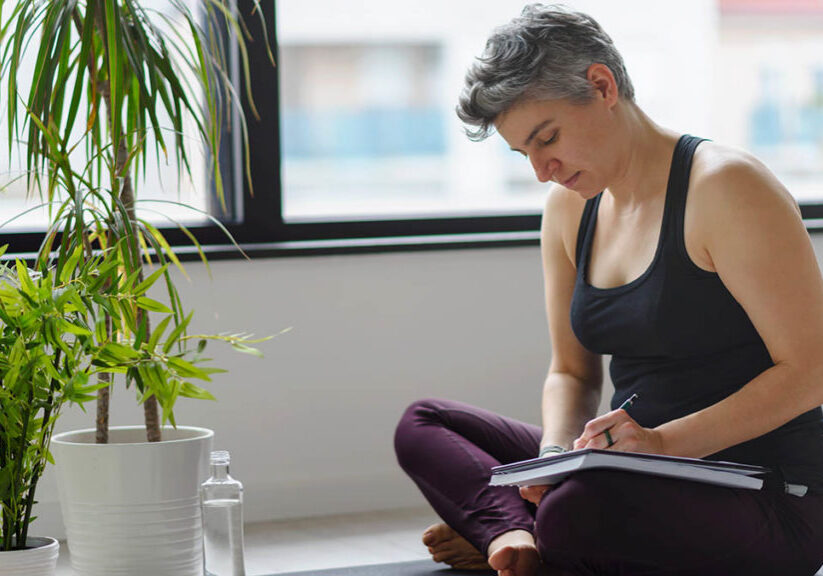 Woman sitting on yoga mat in workout clothes writing in her notebook