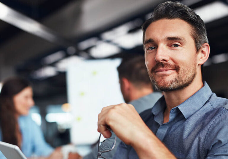 Portrait of a man sitting at a table in an office using a digital tablet with colleagues working in the background.