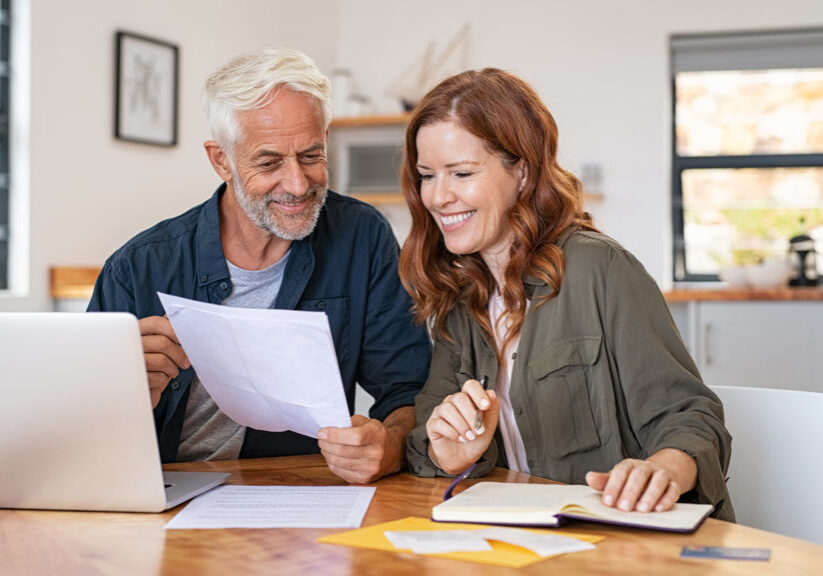 Mature smiling couple sitting and managing expenses at home. Happy senior man and mid woman paying bills and managing budget. Middle aged couple checking accountancy and bills while looking at receipt.