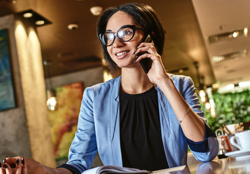 Mixed race businesswoman in blue jacket and glasses talking on phone sitting at cafe table, busy entrepreneur working distantly in coffee house with, papers, talking on mobile, woman making call having lunch in cafeteria